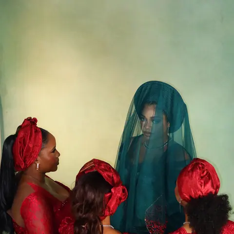 The bride of the CM Chapter wedding under a teal veil, surrounded by three women in red lace dresses and red gele