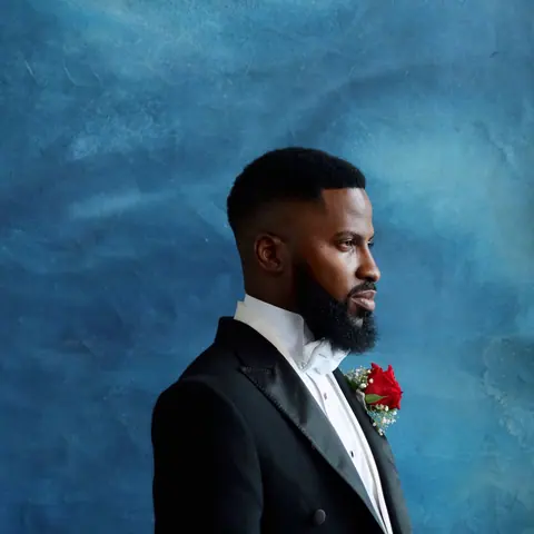The groom of the Faminus wedding in profile, in a black tuxedo with a white bow tie and a red rose, against a deep blue backdrop