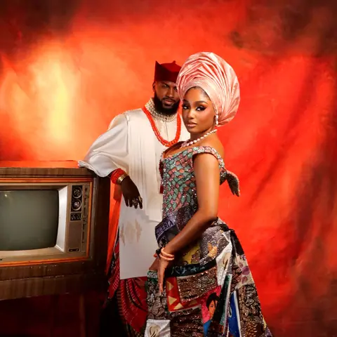 Mr and Mrs Faminus: the groom in white with a red cap and coral beads leans on a vintage television beside the bride in a patchwork gown and salmon gele