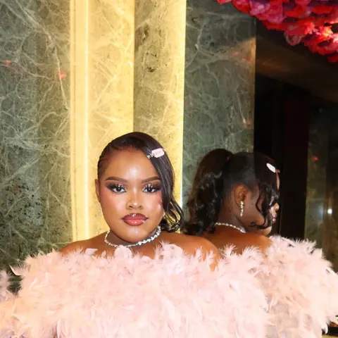 A woman in a blush-pink feather-trimmed sequin dress posing against green marble beneath a ceiling of roses
