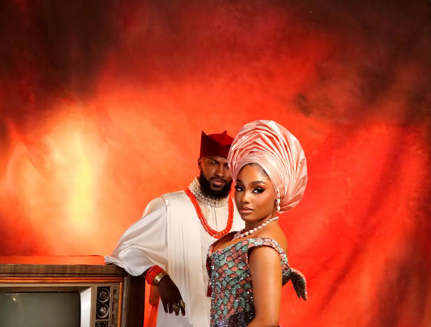 Mr and Mrs Faminus: the groom in white with a red cap and coral beads leans on a vintage television beside the bride in a patchwork gown and salmon gele, against a red-orange painted backdrop