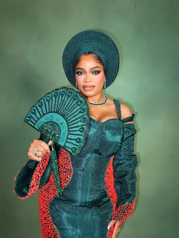 A bride in a bottle-green dress and gele with a green beaded fan