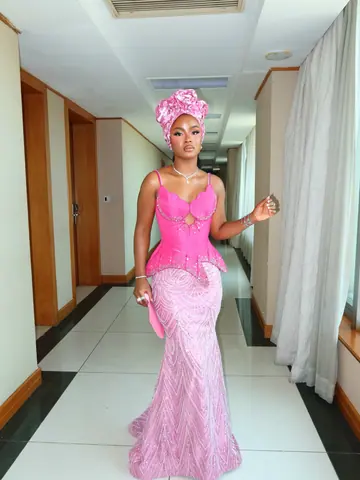 A wedding guest in a hot-pink bodice, pink lace skirt and pink gele in a hotel corridor