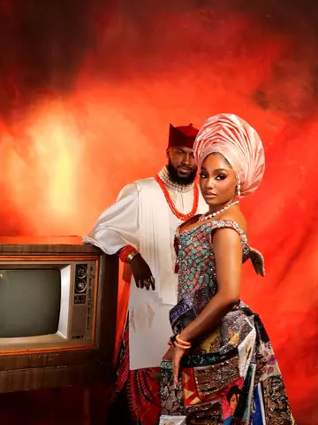 Mr and Mrs Faminus: the groom in white with a red cap and coral beads beside the bride in a patchwork gown and salmon gele, against a red-orange backdrop